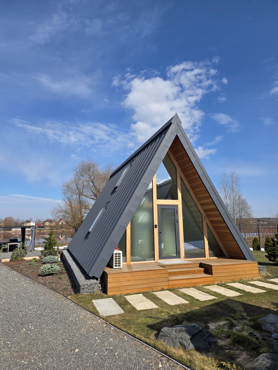 Современный A-frame дом снаружи днём с тёмной крышей, панорамными окнами и деревянной террасой